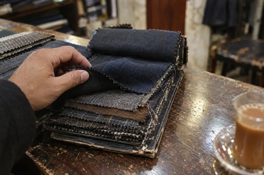 A hand turning the pages of a worn swatch book, with a glass of karak tea on the counter