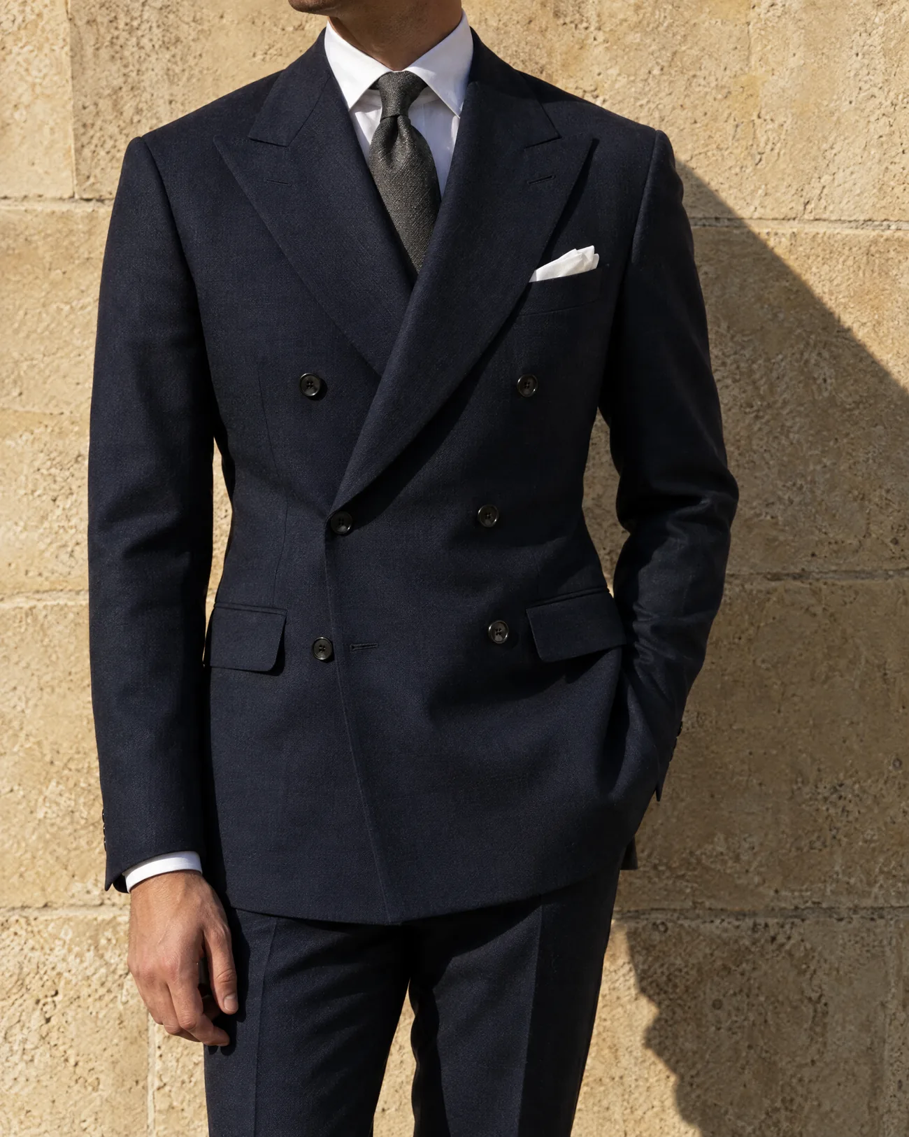A navy double-breasted suit in afternoon sun against a limestone wall